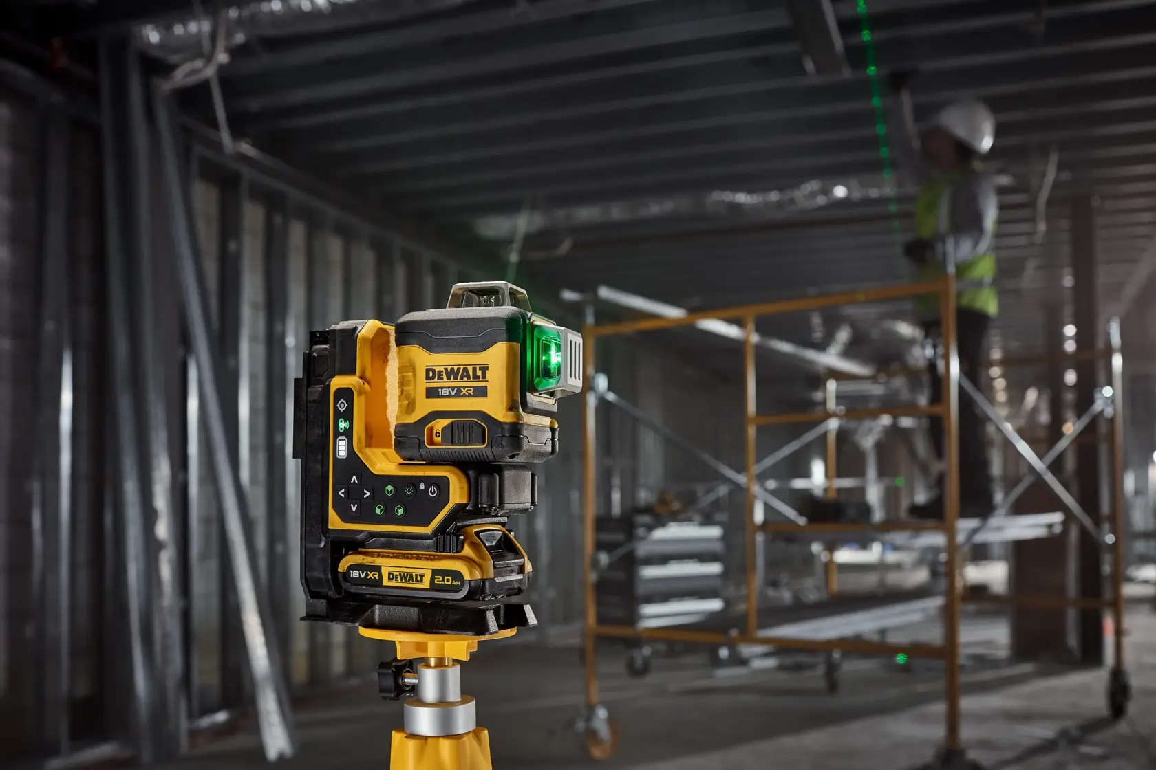 A DEWALT green line laser level is mounted on a metal stud wall in a construction site. The device projects green laser lines for alignment, and a construction worker is visible working in the background.