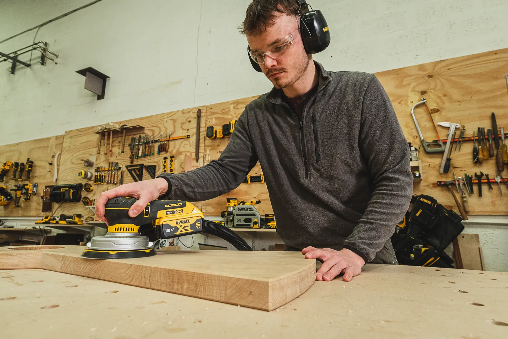 Person sanding wood with a DEWALT DCW230N-XJ sander in a workshop, wearing safety gear.