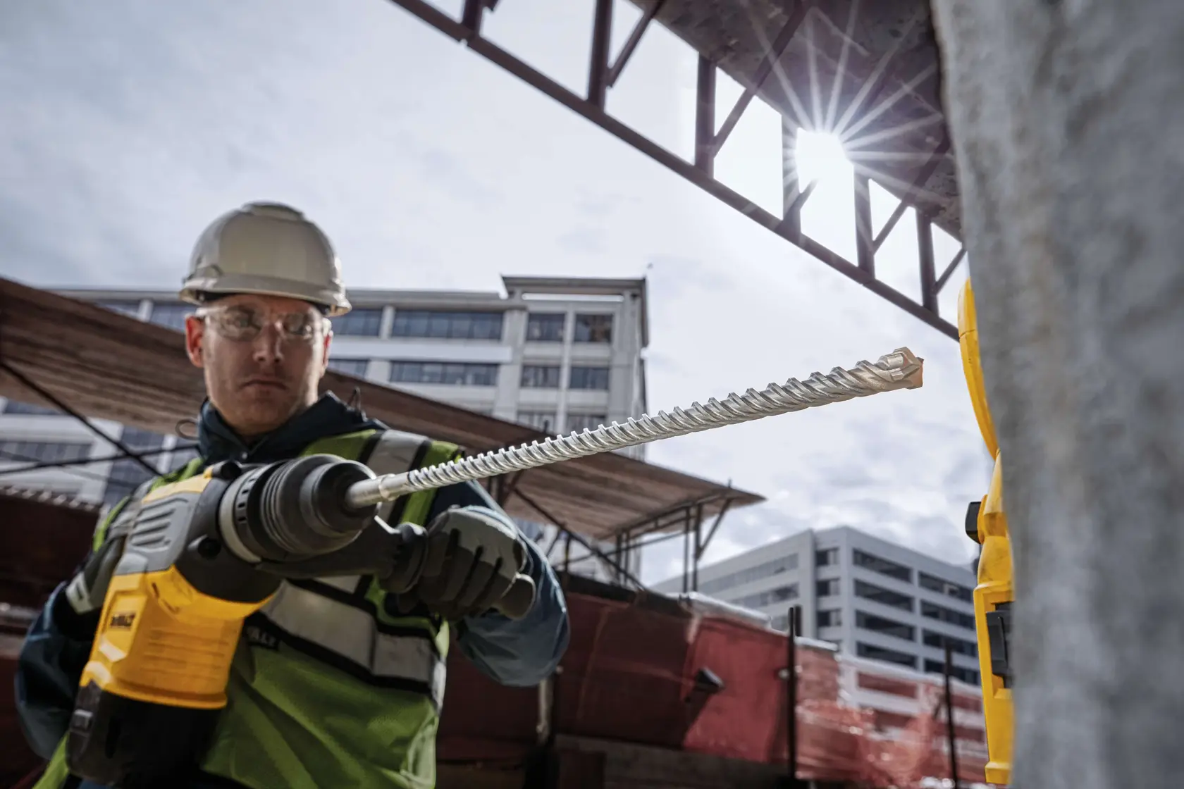 Worker uses a DEWALT drill with DT60825 bit at a construction site with buildings in the background.