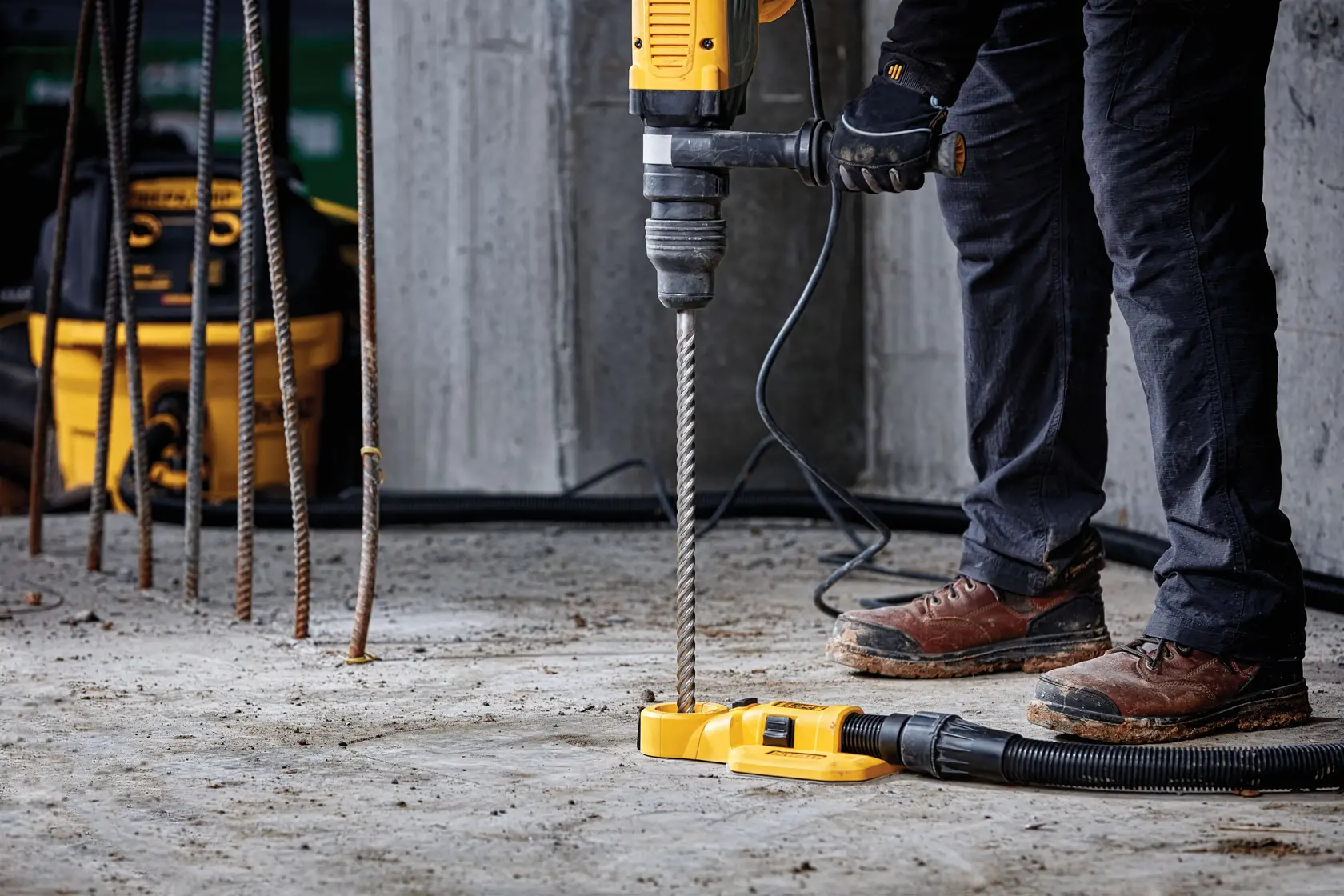 Person using a DEWALT hammer drill with dust extraction on concrete at a construction site. Rebar visible in background.