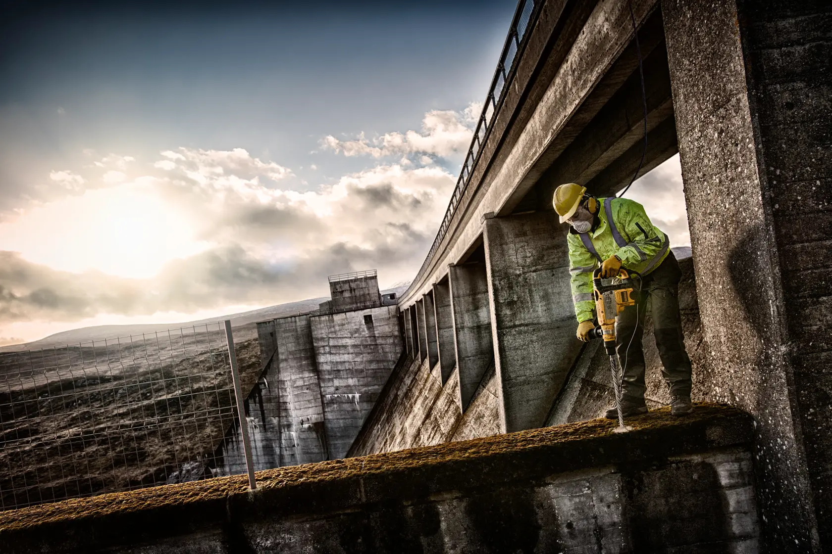 Worker uses DEWALT drill on concrete at construction site with safety gear and bright sky in the background.