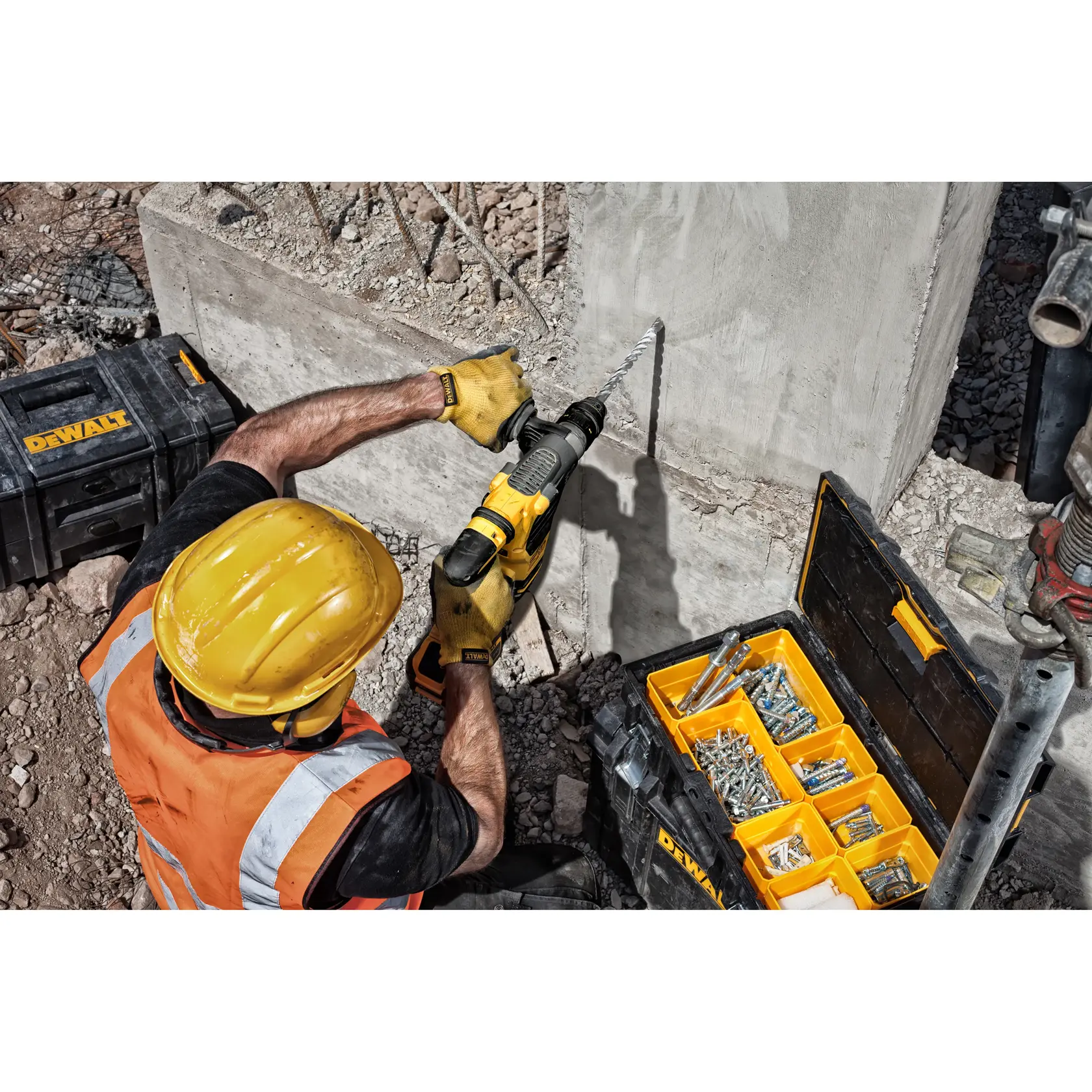 Worker uses a DEWALT drill on concrete, with yellow toolboxes and fasteners organized nearby at a construction site.