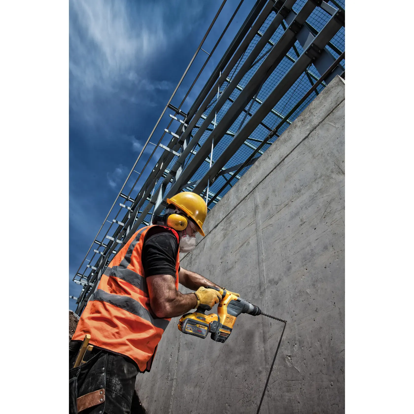 Worker in safety gear uses DEWALT drill on a concrete wall at a construction site under a blue sky.