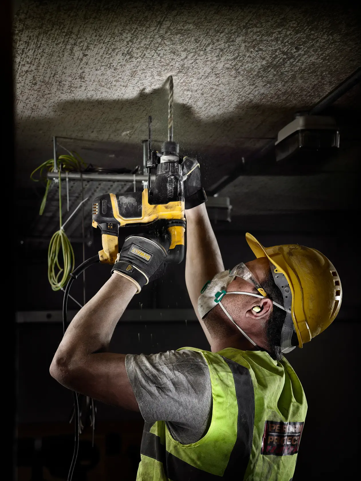 Worker wearing safety gear uses a DEWALT drill to make a hole in a ceiling at a construction site.