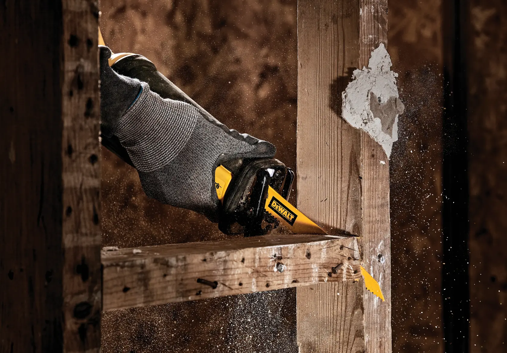 A gloved hand using a DEWALT reciprocating saw blade to cut through a wooden frame, with sawdust and debris flying around.