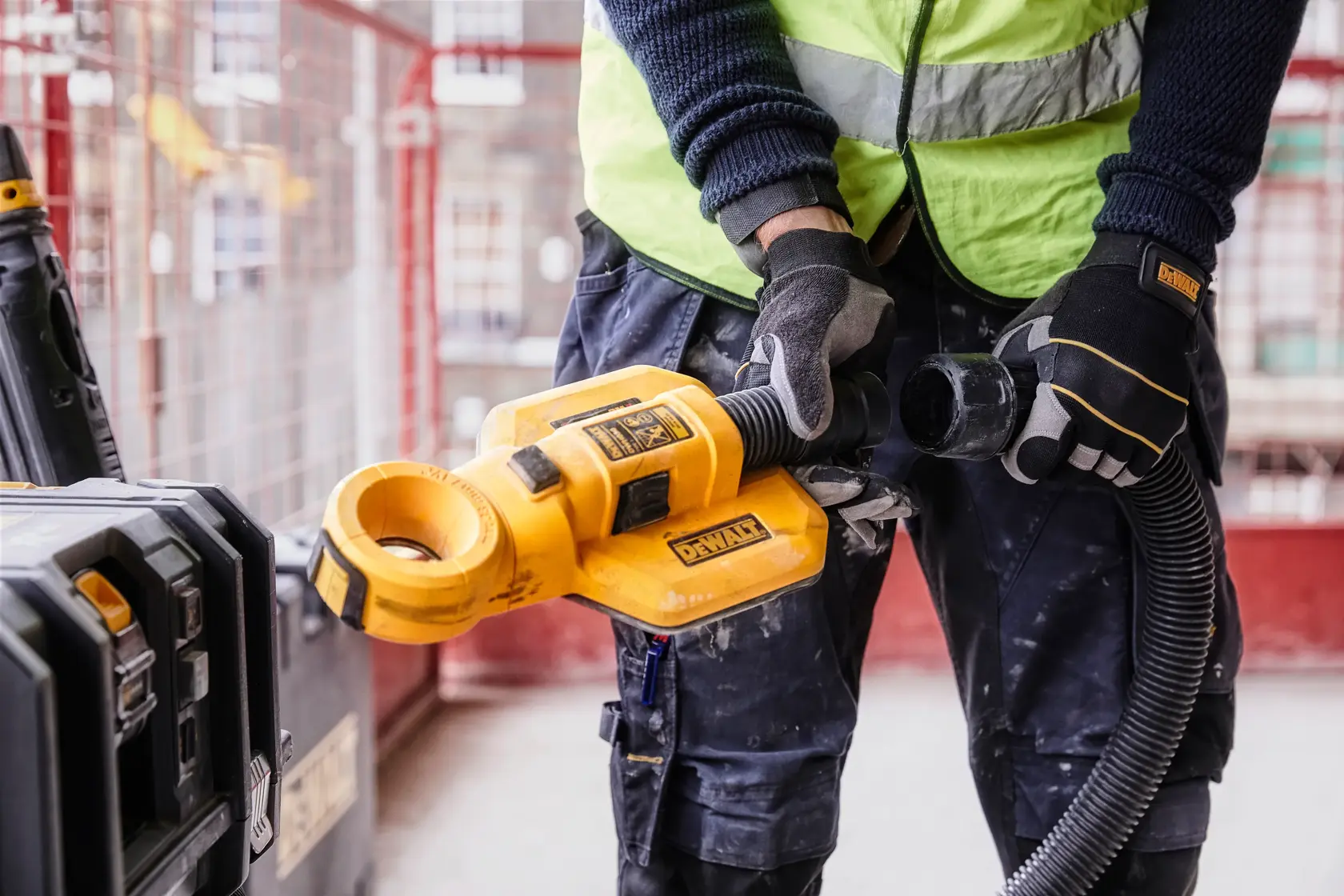 A worker wearing a green safety vest and helmet is using a DEWALT DWH050 dust extraction system attached to a yellow drill, drilling into a concrete ceiling in an industrial setting.