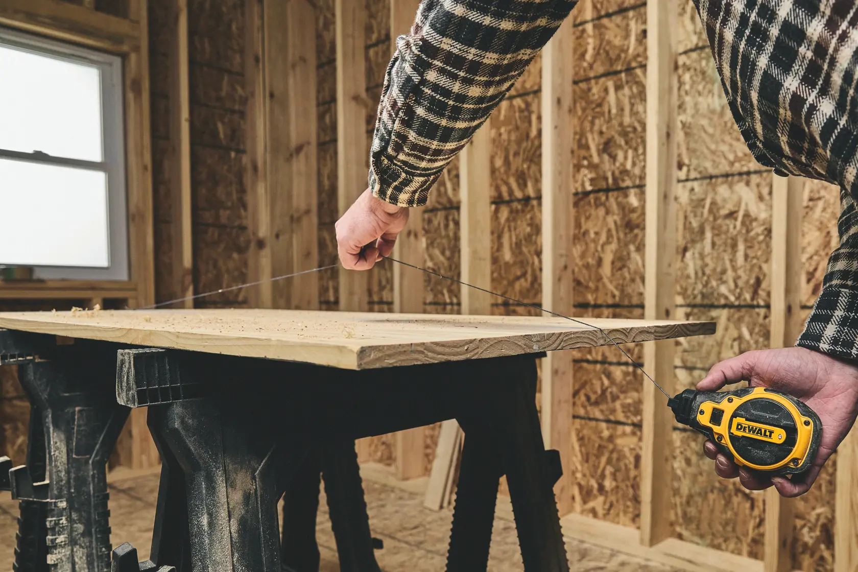 A person using a DEWALT tape measure (model DWHT48333BC) to measure a piece of wood supported by sawhorses in a workshop under construction.