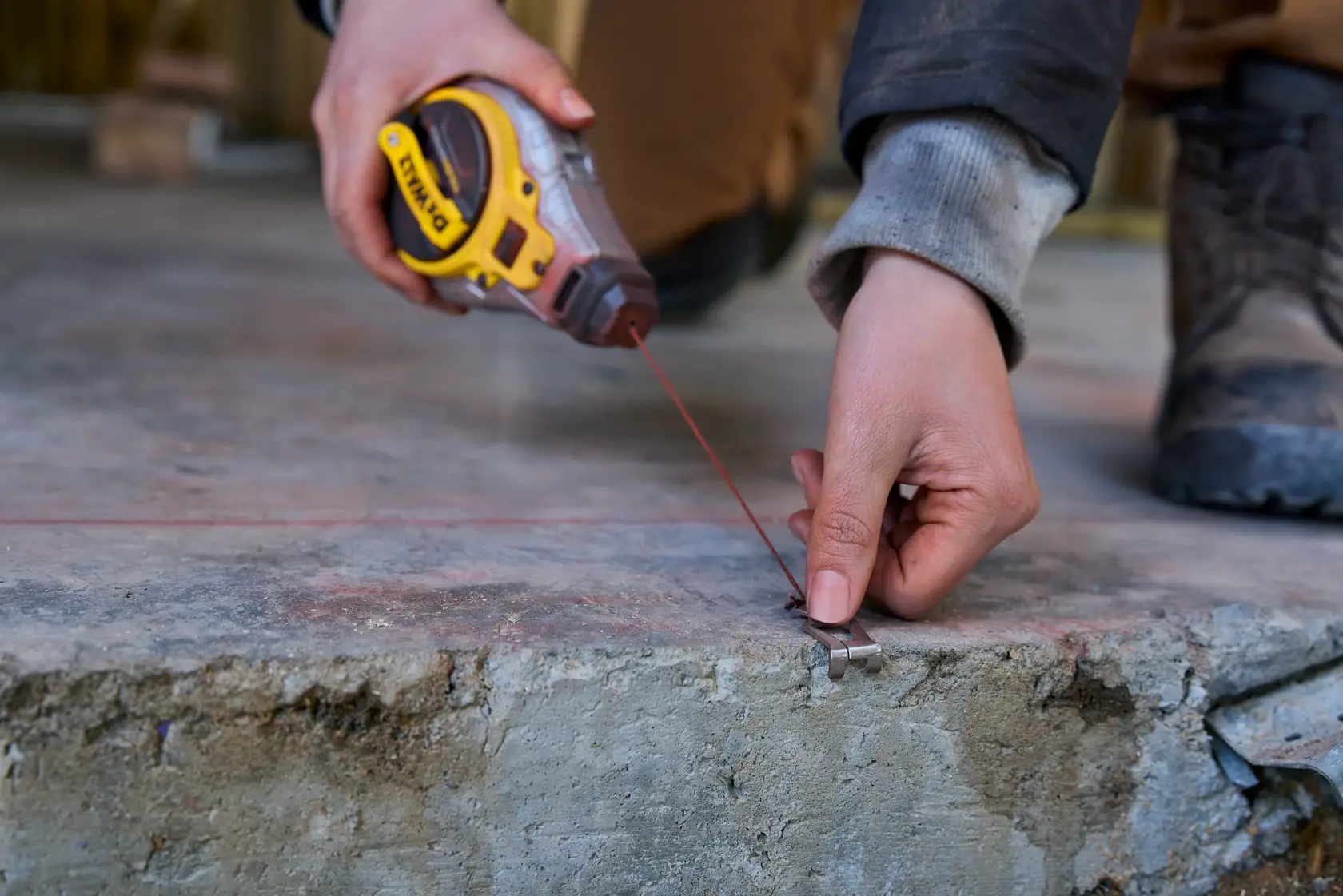 A person uses a DEWALT chalk line tool to mark a straight line on a concrete surface. The person is holding the tool in one hand and securing the chalk line with a metal hook in the other hand.
