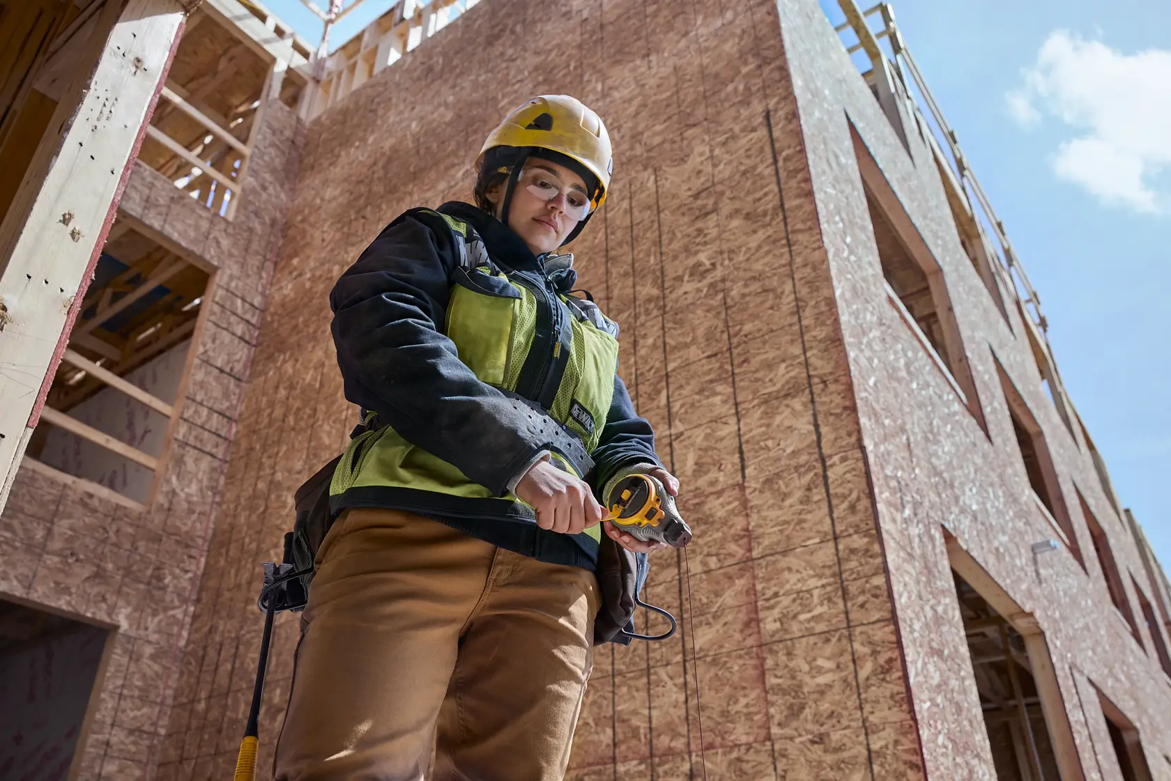A person wearing a yellow safety helmet and green safety vest is standing at a construction site, holding a DEWALT measuring tape. The building structure in the background appears to be under construction.