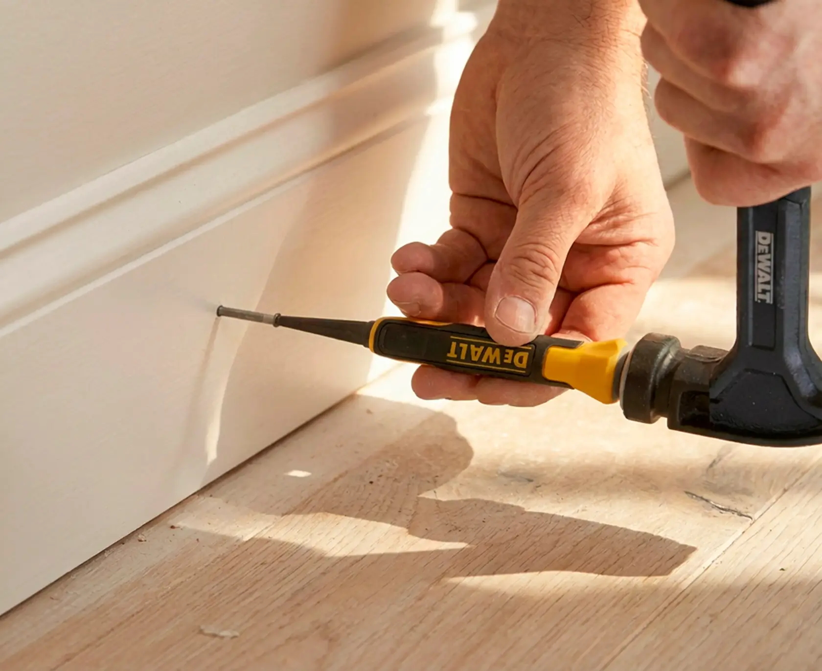 A close-up image showing a person using a DEWALT nail set and hammer to drive a nail into the baseboard of a wall. The surface is a light-colored wood floor.
