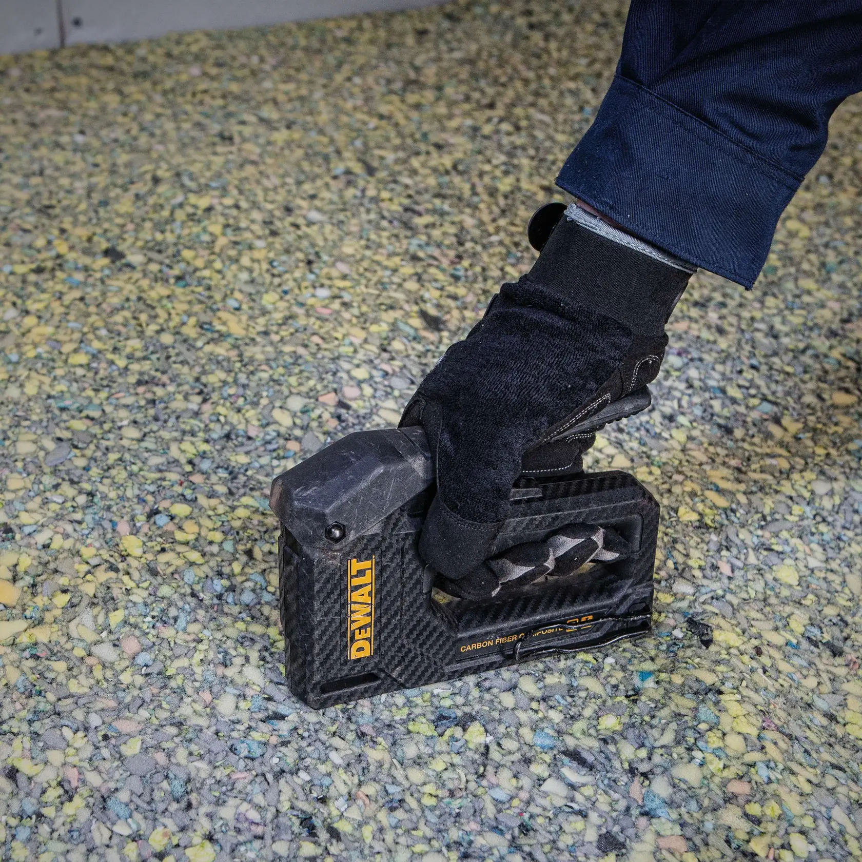 A gloved hand holds a DEWALT carbon fiber stapler (model DWHT80276) against a textured surface, likely for flooring or carpeting installation.