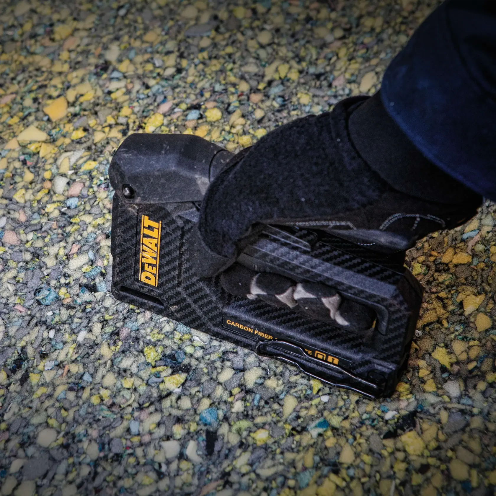 A person wearing a black glove is using a DEWALT carbon fiber hand tool, likely a staple gun, on a textured surface with yellow and multicolored speckles.