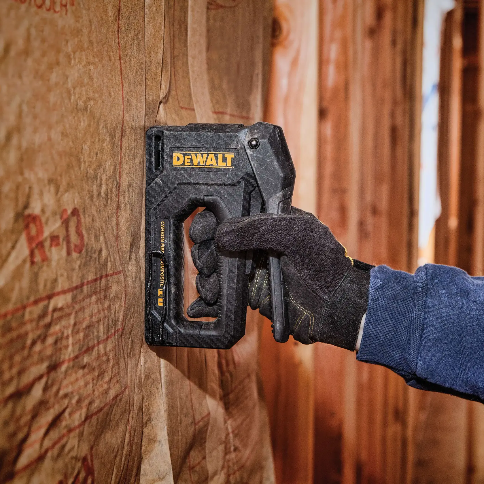A person wearing black gloves using a DEWALT staple gun to secure insulation material to a wooden wall.