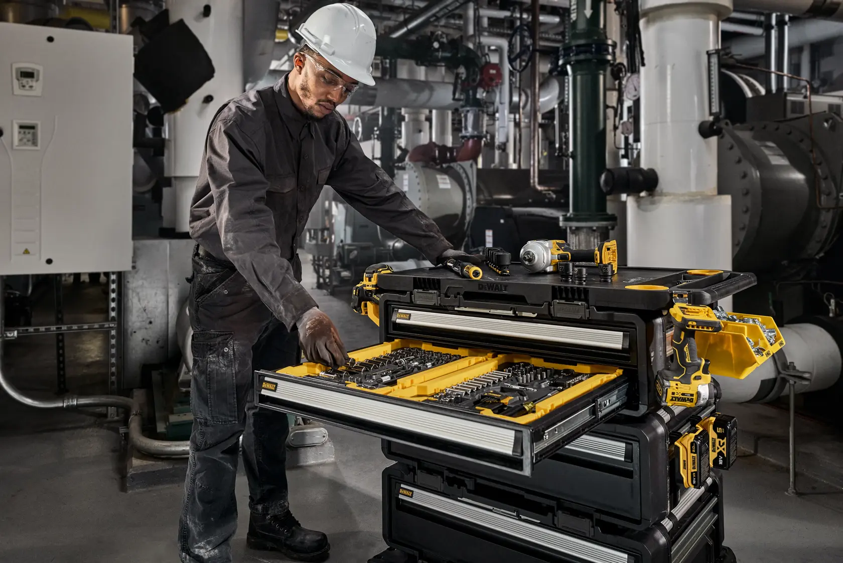 A person wearing protective workwear and a hard hat is using a DEWALT DWST60510-1 tool storage system in an industrial environment. The tool chest is open, displaying organized tools and accessories in multiple drawers.