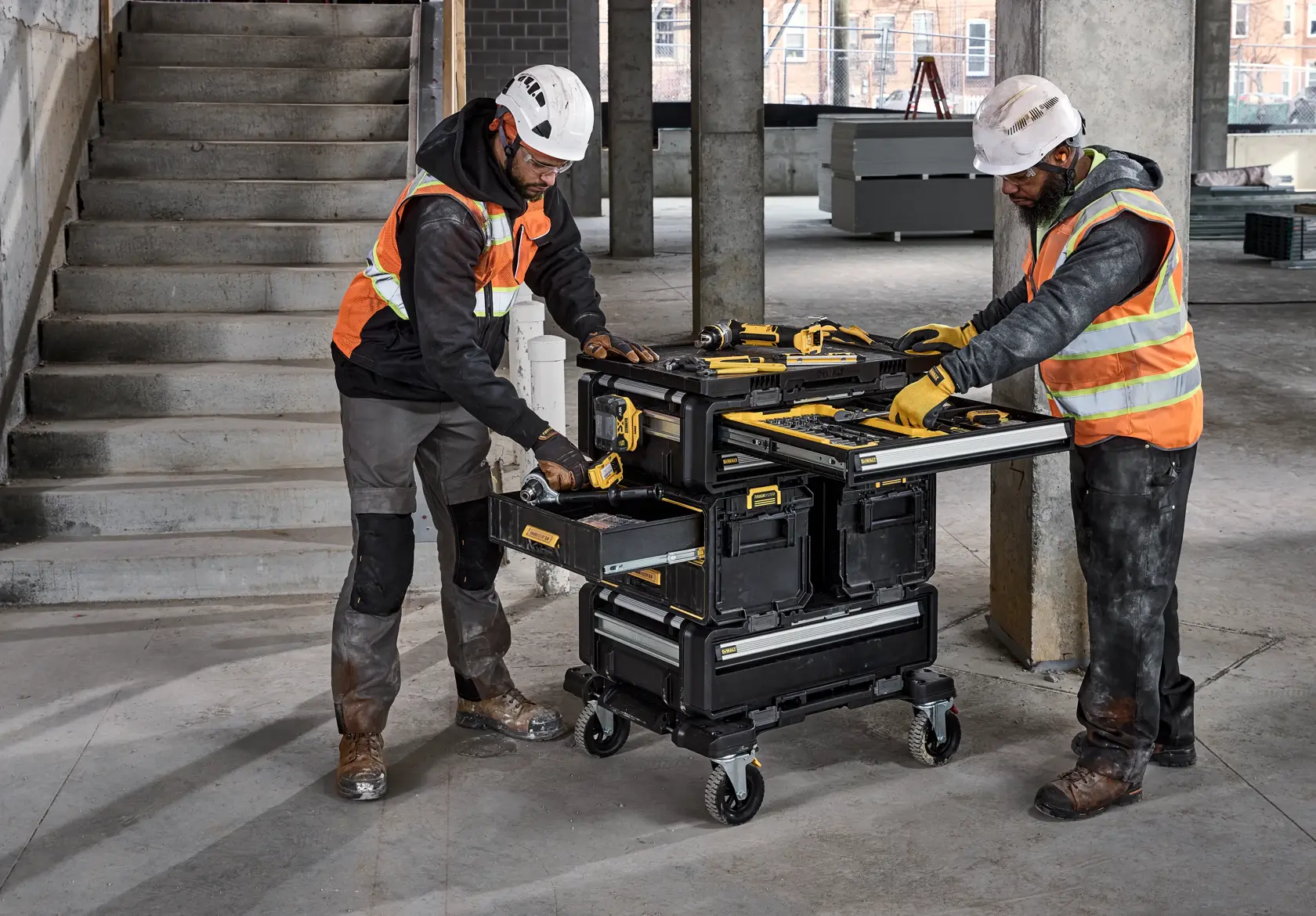 Two construction workers wearing safety helmets and reflective vests are using a DEWALT mobile tool chest on wheels at a worksite. The tool chest features multiple compartments and drawers, and is stocked with various DEWALT tools. The workers are actively accessing the tools, with one opening a drawer and the other using the work surface.